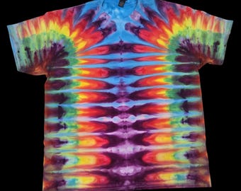 May include: A short-sleeved t-shirt featuring a vibrant tie-dye pattern with a symmetrical design. The colours include bright blue, red, yellow, green, and purple, arranged in repeating vertical bands that radiate from the centre of the garment.