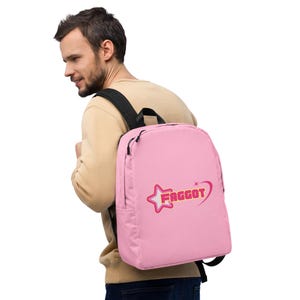 Może przedstawiać: Różowy plecak z czarnymi paskami. Plecak posiada wzór gwiazdy i serca ze słowem "FAGGOT" w stylizowanej czcionce. Plecak jest noszony przez osobę.