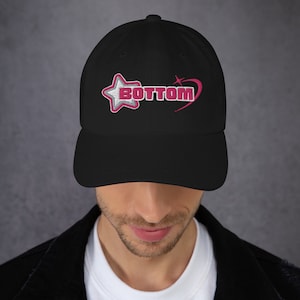 May include: Black baseball cap with the word "BOTTOM" embroidered in white and pink, with a pink star and heart design. The cap has a curved brim and a classic six-panel design.