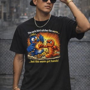 Könnte beinhalten: Schwarzes T-Shirt mit einer Cartoon-Illustration eines blauen Vogels und eines orangefarbenen Wurms in einem Kampf. Der Text auf dem Shirt lautet: "The early bird catches the worm...but this worm got hands!" Das Shirt wird von einer Person getragen.