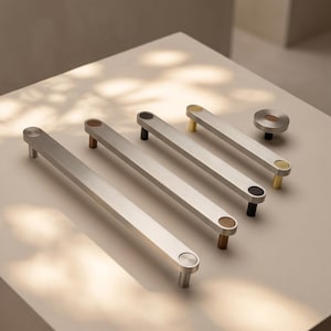 May include: A collection of four elongated silver-toned metal cabinet pulls and one circular knob. Each pull features two circular end caps in varying metallic finishes including gold, bronze, and black, matching the single circular cabinet knob.