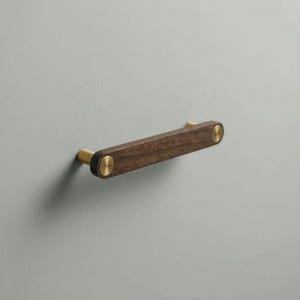 May include: A horizontal drawer pull featuring a dark brown wooden bar with a smooth finish. The bar is secured at both ends by circular, brushed brass metal mounts that connect the handle to the surface, creating a clean and functional design.