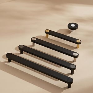 May include: A collection of four matte black cabinet pulls and one circular knob. Each pull features two metallic end caps in varying finishes, including gold, silver, and black. The hardware is arranged in a staggered formation on a flat surface.