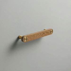 May include: A horizontal cabinet handle featuring a rectangular wooden bar with a visible grain pattern. The handle is secured by two circular brass-toned metal mounts at each end, which connect the wooden bar to the mounting surface.