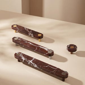 May include: A collection of three elongated drawer pulls and one circular knob, all crafted from deep red marble with white veining. Each piece features metallic accents in gold or silver tones at the mounting points, creating a contrast.