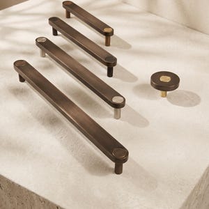 May include: A collection of four dark bronze cabinet pulls of varying lengths and one circular cabinet knob. Each piece features a flat, elongated bar or round face with a contrasting metallic circular inlay at the connection points.