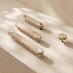 May include: Three elongated rectangular cabinet pulls and one circular cabinet knob made of beige stone with a natural mottled texture. Each piece features metallic hardware accents in silver, gold, and bronze tones at the mounting points.