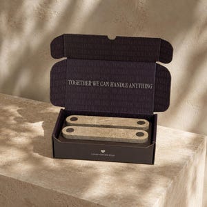 May include: A dark brown cardboard box containing two rectangular beige stone handles with black circular accents. The interior lid features the text TOGETHER WE CAN HANDLE ANYTHING, while the front exterior displays the text Love Handle Duo.