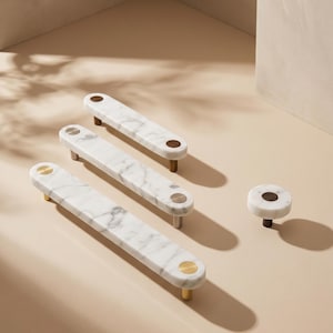 May include: Three elongated white marble cabinet pulls with grey veining and metallic circular accents, alongside one matching round marble knob. Each piece features metallic mounting posts in varying finishes including brass and silver tones.