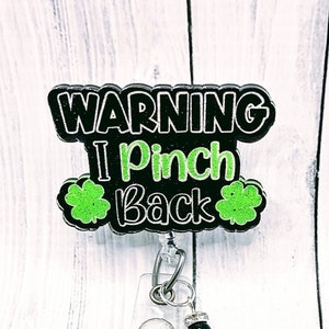 May include: A black and green badge reel with the text "WARNING I Pinch Back" and shamrock accents. The badge reel has a clear plastic holder, a silver retractable cord, and a decorative charm.