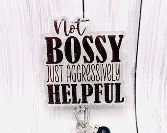 May include: A clear badge holder with a black and silver glitter background. The text on the badge holder reads "Not Bossy Just Aggressively Helpful". The badge holder is attached to a silver retractable badge reel with a black glitter charm.
