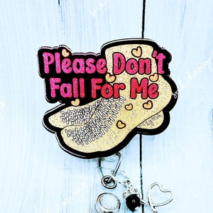 May include: A gold glitter and pink glitter shoe-shaped badge holder with the text "Please Don't Fall For Me". The badge holder is attached to a clear retractable badge reel with a silver heart charm and a black rhinestone charm.
