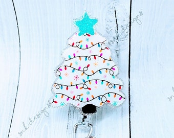 Könnte beinhalten: Ein weißer, weihnachtsbaumförmiger, einziehbarer Ausweishalter mit einem blauen Stern oben und bunten Lichtern. Der Halter hat einen durchsichtigen Plastik-Ausweishalter und eine silberne Kette mit zwei glitzernden Perlen.