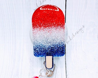 May include: Red, white, and blue glitter popsicle charm with a clear badge holder and a silver retractable badge reel. The charm is attached to the reel with a silver chain and has a small red and blue bead.
