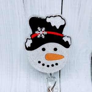 May include: A white glitter snowman with a black top hat, red trim, and a snowflake on top. The snowman has a black mouth, orange nose, and black eyes. The snowman is attached to a clear plastic retractable badge holder with a silver clip and a silver glitter button.