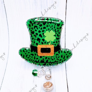 May include: A green and black leopard print badge reel in the shape of a leprechaun hat with a gold glitter buckle and a green shamrock. The badge reel has a clear plastic base with a retractable cord and a decorative bead.