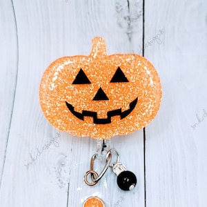 May include: Orange glitter pumpkin with a black jack-o'-lantern face on a retractable badge reel with a black bead and a smaller orange glitter pumpkin charm.