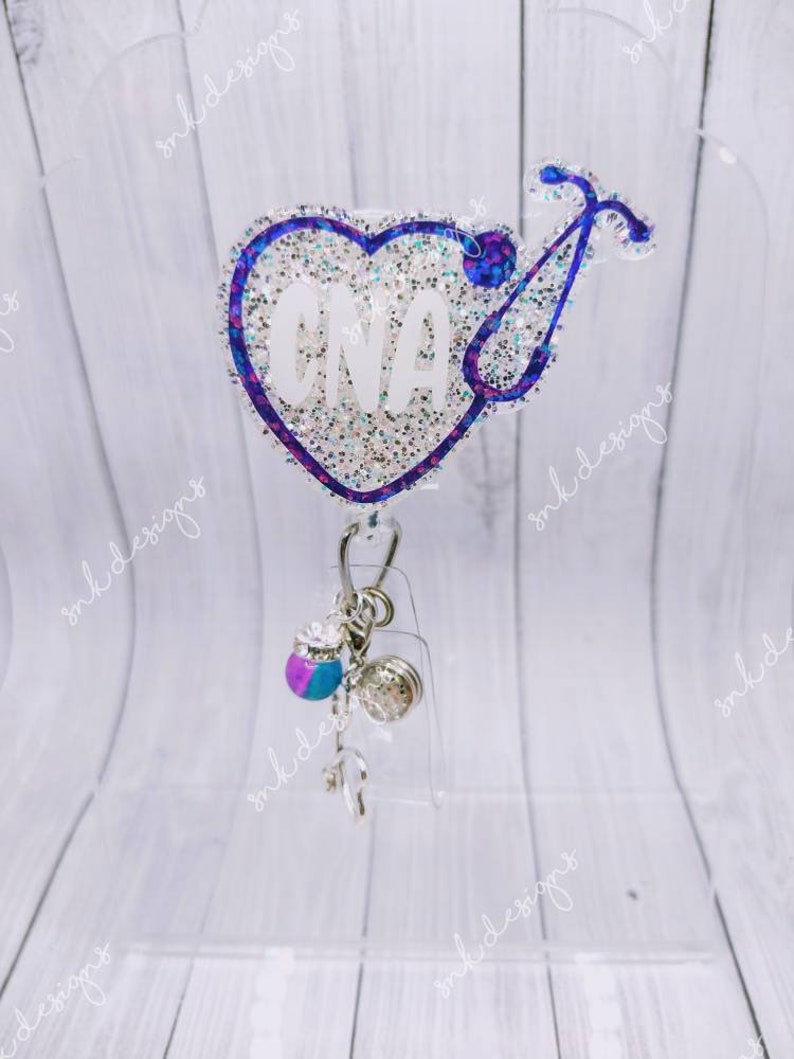 Peut inclure: Un porte-badge en forme de c&oelig;ur scintillant bleu et blanc avec un st&eacute;thoscope et le texte "CNA" en blanc. Le porte-badge est dot&eacute; d'une cha&icirc;ne argent&eacute;e avec un charme bleu et violet et un charme argent&eacute;.