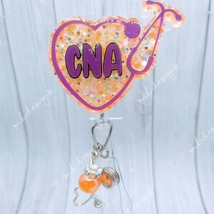 Peut inclure: Un porte-badge en forme de c&oelig;ur violet avec des paillettes et le texte "CNA" &agrave; l'int&eacute;rieur. Un st&eacute;thoscope se trouve sur le c&ocirc;t&eacute; droit du c&oelig;ur. Un porte-badge r&eacute;tractable transparent avec une perle orange et blanche en spirale et un charme en forme de c&oelig;ur argent&eacute; est attach&eacute; au porte-badge.