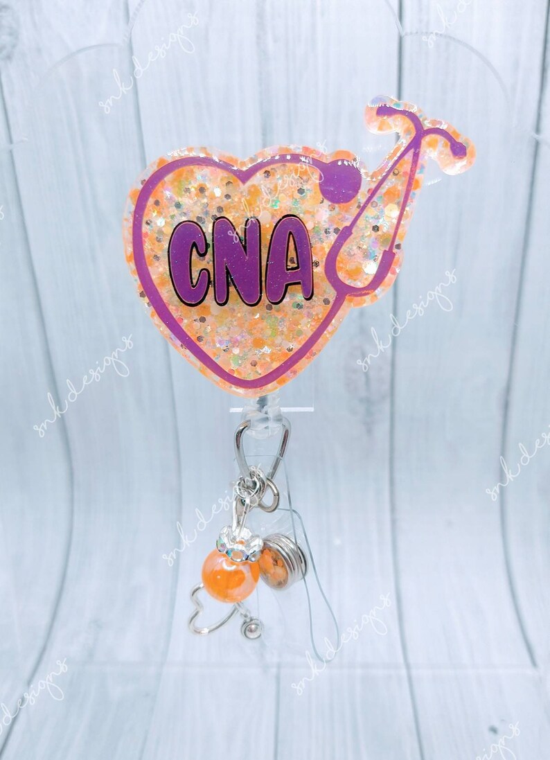 Peut inclure: Un porte-badge en forme de c&oelig;ur violet avec des paillettes et le texte "CNA" &agrave; l'int&eacute;rieur. Un st&eacute;thoscope se trouve sur le c&ocirc;t&eacute; droit du c&oelig;ur. Un porte-badge r&eacute;tractable transparent avec une perle orange et blanche en spirale et un charme en forme de c&oelig;ur argent&eacute; est attach&eacute; au porte-badge.