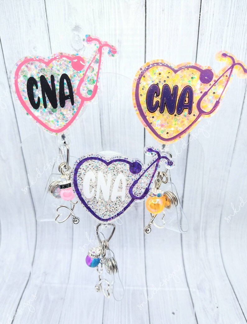 Peut inclure: Trois porte-badges en forme de c&oelig;ur avec des paillettes et le texte "CNA" sur chacun. Les porte-badges sont attach&eacute;s &agrave; des enrouleurs de badge r&eacute;tractables avec diff&eacute;rents breloques.