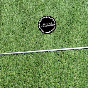 May include: A silver metal tent peg with a hook on one end and a loop on the other end. The peg is lying on a green artificial grass surface.