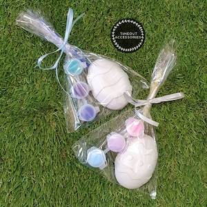 May include: Two white ceramic Easter eggs with paint pots and brushes in clear plastic bags. The bags are tied with blue and white ribbon. The text "TIMEOUT ACCESSORIES" is on a white circle with a green wreath around it.