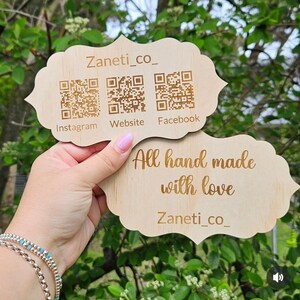 May include: Two wooden signs with a decorative edge are held up. The top sign has the text "Zaneti_co_" and QR codes for Instagram, Website, and Facebook. The bottom sign reads "All hand made with love" and "Zaneti_co_".