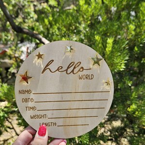 May include: A circular wooden sign with the words "hello WORLD" and star-shaped cutouts. The sign has spaces to write in the baby's name, date, time, weight, and length. The sign is held in front of a green leafy background.