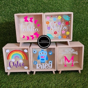 May include: Five wooden shadow boxes with clear glass fronts and colourful designs. The boxes are on small wooden legs. The designs include butterflies, flowers, a rainbow, a monster, and a crown. The boxes are personalised with the names Tahlia, Skyla, Chloe, Riley, and Madison.