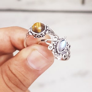 May include: Two silver rings with ornate designs. One ring features a round, brown tiger's eye gemstone. The other ring features a teardrop-shaped, iridescent moonstone.