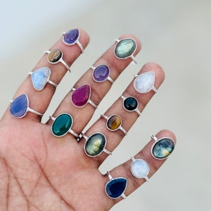 May include: A collection of 14 silver rings with various gemstone settings. The rings feature a variety of colors, including blue, green, red, purple, black, and brown. The gemstones are all in a teardrop shape.