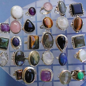Peut inclure: Une collection de 20 bagues en argent avec différentes pierres précieuses. Les pierres comprennent l'améthyste, l'œil de tigre, la pierre de lune, le labradorite, et plus encore. Les bagues sont présentées sur un fond blanc et bleu.