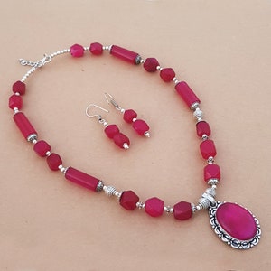 Puede incluir: Un collar y pendientes de piedras preciosas rosas con detalles plateados. El collar presenta un gran colgante ovalado con una piedra preciosa rosa y un marco plateado. Los pendientes son simples aros plateados con una piedra preciosa rosa.