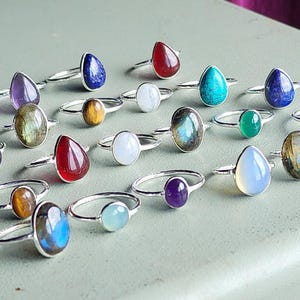 Op de afbeelding: Een verzameling zilveren ringen met verschillende edelsteen bezettingen. De ringen hebben een verscheidenheid aan kleuren en vormen, waaronder rond, ovaal en druppelvormig. De edelstenen zijn gezet in zilver en worden omringd door een zilveren band.