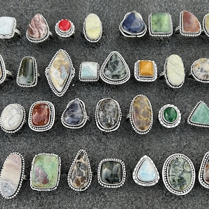 May include: A collection of 25 silver rings with various colorful gemstone inlays. The rings feature a variety of shapes, including oval, square, and triangular. The gemstones are set in silver bezels and have a polished finish.