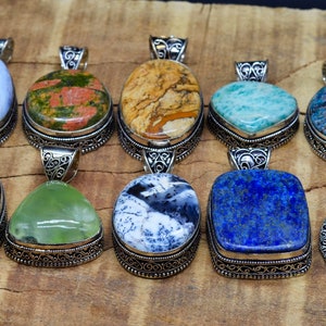 May include: Ten gemstone pendants with silver-coloured metal settings. The pendants showcase diverse stones, such as blue lace agate and turquoise. The stones are oval, teardrop, and square-shaped, with detailed metalwork around the edges. The pendants are displayed on a wooden surface.