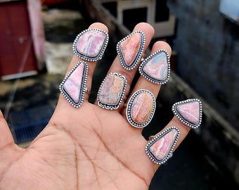 May include: A set of eight silver rings with pink and white agate stones. The rings are all different shapes and sizes, and they are arranged in a row on a hand.