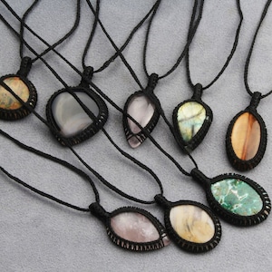 Puede incluir: Una colección de ocho collares con colgantes de piedras preciosas de diferentes colores. Cada colgante está envuelto en un cordón negro y tiene una forma y un patrón únicos. Los collares se muestran sobre una superficie gris claro.