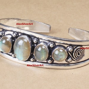 Puede incluir: Pulsera de brazalete de plata con cinco piedras ovaladas de labradorita engastadas en una banda de plata. La banda tiene un diseño decorativo de espiral y pequeñas cuentas de plata.