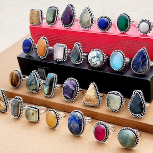 Op de afbeelding: Een verzameling zilveren ringen met diverse kleurrijke edelstenen, waaronder amethist en lapis lazuli. De ringen hebben sierlijke zilveren zettingen en worden gepresenteerd op een rode doos en een bruin oppervlak.