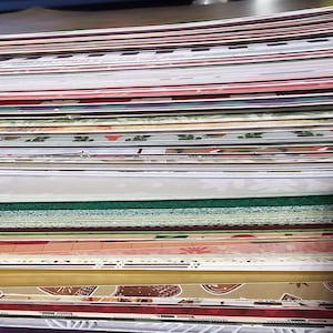 May include: A stack of decorative paper sheets with various patterns and colors. The papers include designs with red, green, and gold accents, suitable for crafts or scrapbooking. The stack is viewed from a low angle, showing the layered arrangement.