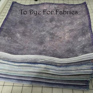May include: A stack of fabric squares with a textured, mottled appearance in shades of blue, gray, and purple. The edges are neatly finished with a purple stitch. The text "To Dye For Fabrics" is visible on the top square.