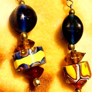 May include: A pair of handmade earrings featuring a dark blue oval bead, followed by amber-colored beads, a yellow and blue patterned bead, and a red bead. The earrings are suspended from a silver-colored hook.