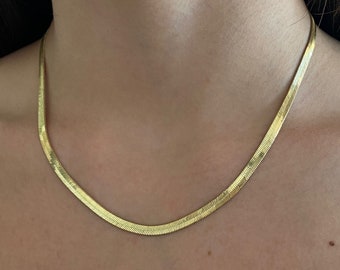 Könnte beinhalten: Eine goldfarbene Halskette mit Fischgrätenmuster. Die Halskette ist eine flache, flexible Kette mit einem dezenten, strukturierten Muster. Die Halskette wird um den Hals getragen und liegt auf der Brust.