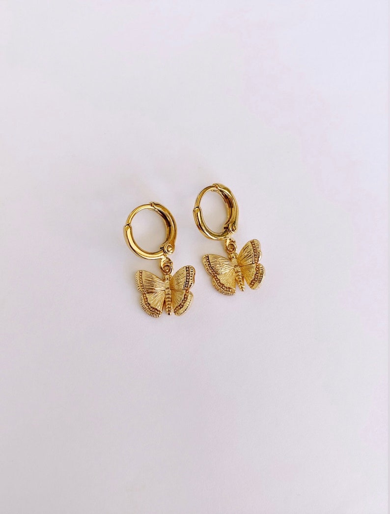 May include: A pair of gold hoop earrings with a dangling butterfly charm. The butterfly charm is detailed with textured wings.