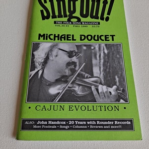 Könnte beinhalten: Ein grünes Magazincover mit dem Titel "Sing Out!" The Folk Song Magazine, Vol. 35 #3, Herbst 1990, 3,75 $. Das Cover zeigt ein Schwarzweißfoto von Michael Doucet mit dem Text "Cajun Evolution".