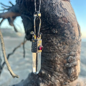 May include: A pendant necklace featuring a clear, pointed crystal suspended from a textured, dark metal cap. The cap is accented with a small, round, deep red gemstone and gold-toned flecks, hanging from a twisted silver-toned metal chain.