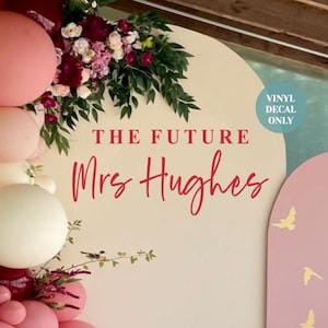 May include: A pink and white balloon arch with greenery and flowers. The background is a light beige wall with a pink and beige arch. The text "THE FUTURE Mrs Hughes" is written in red on the wall. A small blue circle with the text "VINYL DECAL ONLY" is in the top right corner of the image.