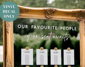 Op de afbeelding: Een grote, sierlijke goudkleurige lijst met een doorzichtige acrylplaat voorzien van witte vinyltekst "OUR FAVOURITE PEOPLE" en "your seat awaits". Twaalf witte tafelkaartjes zijn met gouden clips aan het doorzichtige oppervlak bevestigd.